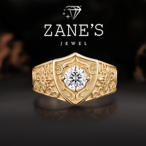 May include: A gold ring with a central, round, clear gemstone. The ring has a shield-like design with floral and scrollwork patterns. The text "ZANE'S JEWEL" is above the ring.