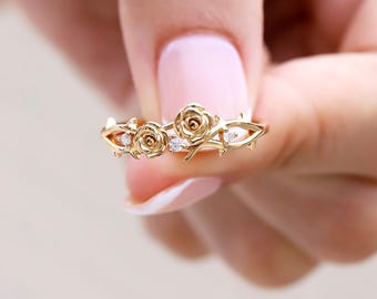 Op de afbeelding: Een gouden ring met twee gebeeldhouwde rozenbloesems en doornige rankdetails langs de band. Het ontwerp bevat drie kleine, heldere, rond geslepen edelstenen die tussen de rozen en de metalen rankelementen zijn geplaatst voor een bloemmotief.