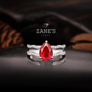 Puede incluir: Un anillo de rubí en forma de pera engastado en una banda de plata con una doble fila de diamantes pavé. El anillo está sobre un fondo oscuro con el texto "ZANE'S JEWEL" encima.