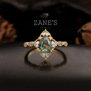 May include: A gold ring featuring a pear-shaped green moss agate gemstone, surrounded by smaller clear gemstones. The ring has a delicate, intricate design and is set against a black background. Zane's Jewel.