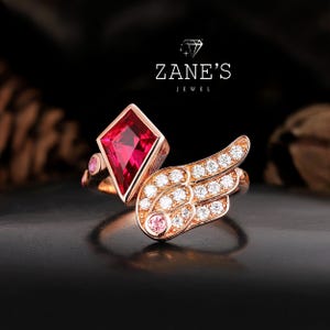 May include: A rose gold ring featuring a large, red, diamond-shaped gemstone and a wing design encrusted with small, clear gemstones. The ring also includes a small pink gemstone. The text "ZANE'S JEWEL" is visible.