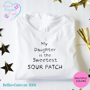以下が含まれることがあります： 黒い文字で「My Daughter is the Sweetest SOUR PATCH」と書かれた白いBella+Canvas 3001 Tシャツ。 シャツはクルーネックで折りたたまれています。 画像にはロゴと「MULTIPLE COLORS」と書かれたピンク色の円も含まれています。