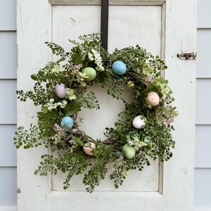 May include: A green and white Easter wreath with pastel colored eggs. The wreath is made of faux greenery and is hanging on a white door.
