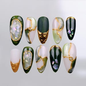 May include: Ten green and white almond-shaped fake nails with gold accents. The nails have a variety of designs, including swirls, geometric shapes, and glitter.