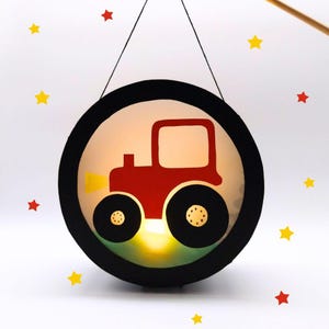 Może przedstawiać: Okrągła latarnia z czarną ramą i czerwonym wzorem traktora. Traktor ma czarne koła i żółty reflektor. Latarnia wisi na czarnym sznurku, z żółtymi i czerwonymi gwiazdami.