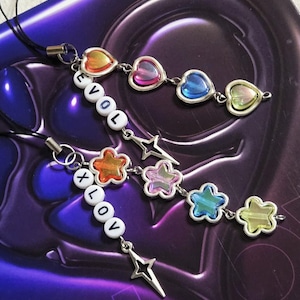 May include: Two beaded phone charms with colorful heart and star charms. One charm spells "EVOL" and the other spells "XLOV" with white letter beads. The charms have silver-toned metal accents and black cords.