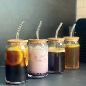 Könnte beinhalten: Vier Glasgefäße mit Bambusdeckeln und Glasstrohhalmen, gefüllt mit verschiedenen Getränken. Die Gefäße stehen auf einer grauen Oberfläche. Die Getränke umfassen Eiskaffee mit einer Zitronenscheibe, ein rosafarbenes Eisgetränk, einen dunklen Eiskaffee und einen hellbraunen Eistee.