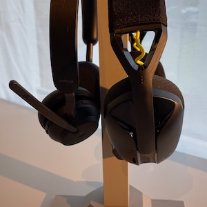 Puede incluir: Dos auriculares negros con diseños diferentes están colgados de un soporte beige. El soporte tiene una base geométrica. Los auriculares tienen una diadema y almohadillas negras. El auricular izquierdo tiene un micrófono y el texto "Jabra" en la almohadilla. El auricular derecho tiene un acento amarillo en la diadema.