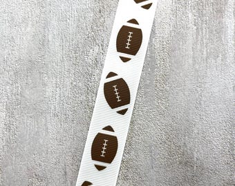 May include: White ribbon featuring a repeating pattern of brown American footballs. Each football is detailed with white stitching. The ribbon is angled across a textured, grey background. Ideal for crafting and sports-themed projects.