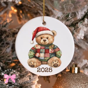 May include: A white ceramic Christmas bauble with a teddy bear wearing a Santa hat and a patchwork sweater. The bear has brown fur and is sitting down. The year "2025" is printed below the bear. A gold string is attached for hanging.