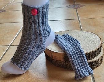 May include: A pair of grey knitted ankle warmers with a pink "Hand Made" tag on one of the warmers. The warmers are on a wooden surface.