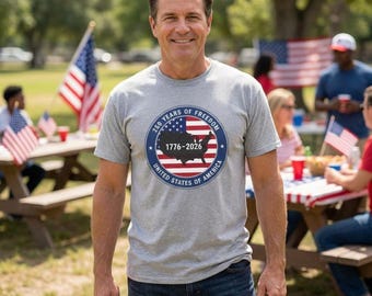 May include: A light grey short-sleeved t-shirt featuring a circular graphic on the chest. The graphic displays a map of the United States, the American flag, and the text 250 YEARS OF FREEDOM 1776-2026 UNITED STATES OF AMERICA in blue and white.