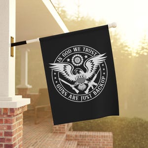 Op de afbeelding: Zwarte vlag voor buiten met een witte afbeelding van een adelaar, geweren en de tekst "IN GOD WE TRUST" en "GUNS ARE JUST BACKUP". De vlag hangt buiten aan een witte paal.