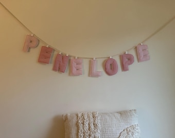 Puede incluir: Una guirnalda con la palabra "PENELOPE" cuelga de una pared crema. Las letras son de tela rosa, algunas con un diseño a rayas. La guirnalda está suspendida de una cuerda fina y clara, con cada letra sujeta por pequeñas pinzas.