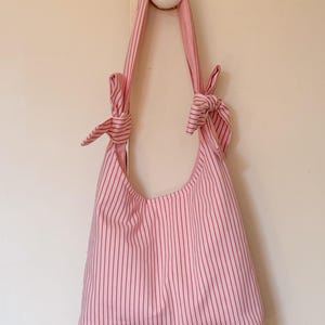 May include: A pink and red striped shoulder bag hanging on a wall. The bag has a rounded shape and is made of fabric with vertical stripes. The straps are tied in knots at the top. The bag is hanging from a white wall.
