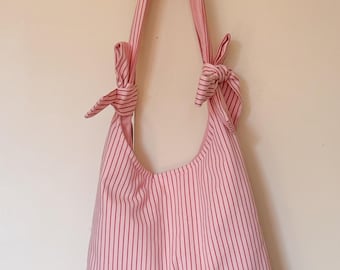 Peut inclure: Sac à bandoulière à rayures roses et rouges suspendu à un mur. Le sac a une forme arrondie et est fait de tissu à rayures verticales. Les sangles sont nouées en haut. Le sac est suspendu à un mur blanc.