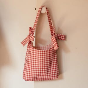 May include: A red and white checked tote bag with a shoulder strap. The strap is tied with bows on each side of the bag. The bag is hanging on a white wall.
