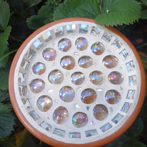 May include: A circular terracotta saucer featuring a mosaic design. The centre is filled with iridescent, round glass cabochons embedded in white grout, while the outer rim is lined with small, rectangular mirror tiles.