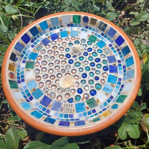 May include: A circular mosaic bird bath with a terracotta rim. The mosaic design incorporates blue, green, and white tiles, with glass beads and seashells at the centre. The bird bath is positioned amidst green grass and leaves.