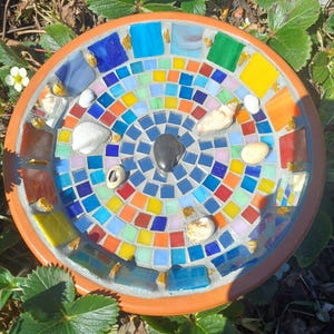 May include: A circular terracotta bird bath featuring a mosaic design made of multicoloured glass tiles in shades of blue, green, yellow, orange, and red. The centre contains a smooth black stone, accented by several small white seashells.