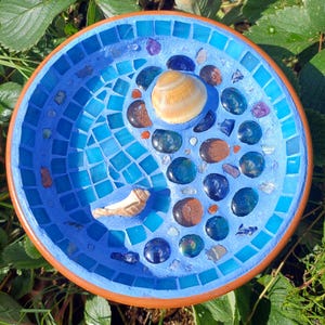May include: A circular, blue mosaic bird bath with a brown rim. The mosaic design incorporates blue tiles, seashells, and colourful stones. The bird bath is situated outdoors amidst green foliage.
