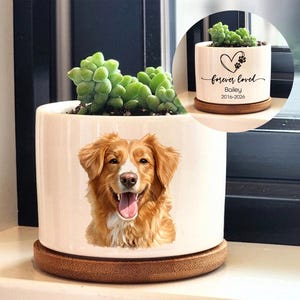 May include: A white ceramic planter featuring a smiling dog portrait, set on a round wooden base. The planter holds a green succulent plant. The inset image shows a similar planter with the text "forever loved" and a pet's name.
