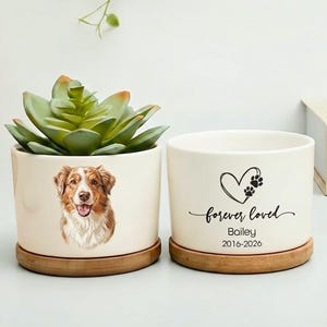 May include: Two white ceramic planters with wooden bases. One features a dog portrait, the other has a heart and paw print design with the text "forever loved" and a name and dates. A succulent plant sits in one.