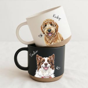 May include: Two ceramic mugs stacked on top of each other. The top mug is white with a brown base and a drawing of a dog named Ruby. The bottom mug is black with a brown base and a drawing of a dog named Sadie.