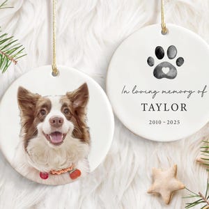 May include: Two white ceramic hanging ornaments. One has a dog portrait, the other a paw print with a heart and the text "In loving memory of TAYLOR 2010-2025". A pet memorial decoration.