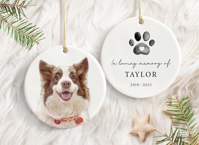 May include: Two white ceramic hanging ornaments. One has a dog portrait, the other a paw print with a heart and the text "In loving memory of TAYLOR 2010-2025". A pet memorial decoration.