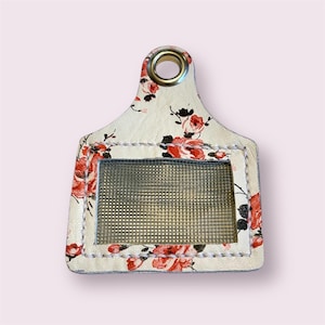 May include: Cream-colored tag with a floral pattern of red roses and black accents. The tag has a rectangular window with a mesh insert and a gold-colored grommet at the top. The edges are stitched with white thread.