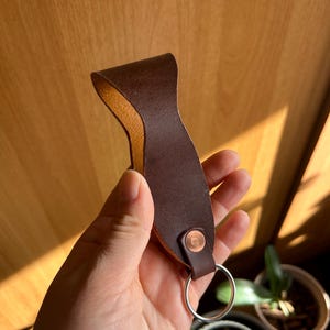 May include: A brown leather key chain with a metal ring. The leather is folded over and stitched together. The key chain has a brown leather loop at the top.
