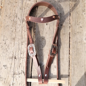 May include: A brown leather horse bridle with heart-shaped accents and decorative silver buckles. The bridle features a wooden bit and is set against a rustic wooden background.
