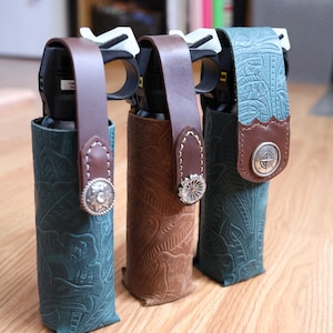 Peut inclure: Trois bombes de poivre logées dans des étuis en cuir gaufré. Deux étuis sont bleu sarcelle avec des motifs floraux, et un est marron avec un motif floral. Chacun dispose d'une sangle en cuir marron fixée par un bouton concho en métal argenté.