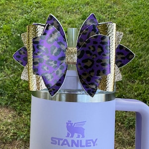 May include: A purple and gold leopard print bow with gold glitter accents is attached to a stainless steel and lavender Stanley travel mug with the Stanley logo.