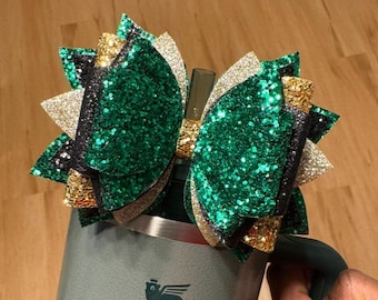 May include: A large green, black, and gold glitter bow with a layered design. The bow is attached to a stainless steel tumbler.