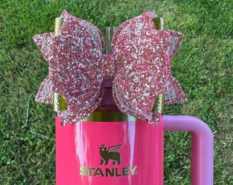 Puede incluir: Taza de viaje rosa Stanley con un lazo de purpurina dorada en la tapa. La taza tiene un logotipo negro con un oso y la palabra "STANLEY".