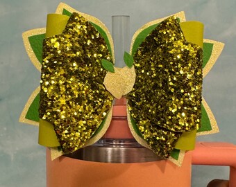 Op de afbeelding: Een groene en gouden glitter strik met een groen bladontwerp zit bovenop een perzikkleurige tumbler met een deksel van roestvrij staal. De tumbler heeft een logo van een kroon en een varken aan de voorkant.