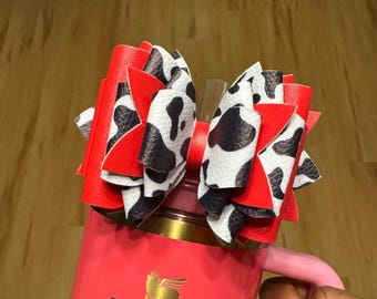 Op de afbeelding: Een handgemaakte haarstrik met een koeienprintontwerp met zwarte en witte vlekken, gelaagd over helderrood materiaal. De strik is bevestigd aan een roze beker met een gouden rand en een roze handvat.
