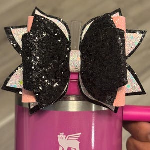 May include: A black glitter bow with white and pink accents is attached to a pink Stanley tumbler. The bow is made of layered ribbons and sits atop the tumbler's straw. The tumbler has a handle and the Stanley logo.