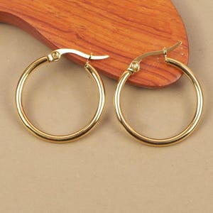 Peut inclure: Deux boucles d'oreilles créoles dorées. Les boucles d'oreilles sont rondes et présentent un design simple et classique. Elles sont présentées sur une surface en bois avec un fond neutre. Les boucles d'oreilles sont en métal brillant et réfléchissant.