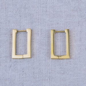 Könnte beinhalten: Zwei goldfarbene, rechteckige Creolen. Die Ohrringe haben ein schlichtes, geometrisches Design mit einem Klappverschluss. Sie werden auf einer hellgrauen, strukturierten Oberfläche präsentiert.