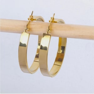 Peut inclure: Une paire de boucles d'oreilles créoles dorées avec un design plat et rectangulaire. Les boucles d'oreilles sont présentées sur une cheville en bois sur fond blanc. Les boucles d'oreilles ont une surface brillante et réfléchissante.