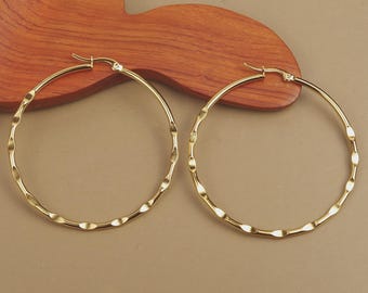 Könnte beinhalten: Ein Paar goldfarbene Creolen mit einem strukturierten, wellenförmigen Design. Die Ohrringe sind kreisförmig und haben einen einfachen Verschluss. Der Schmuck wird auf einer Holzoberfläche präsentiert.