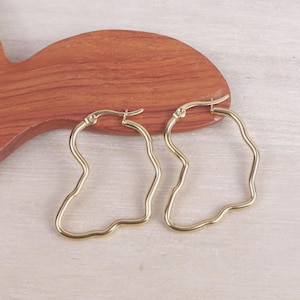Peut inclure: Une paire de boucles d'oreilles créoles dorées avec un contour irrégulier et ondulé. Les boucles d'oreilles sont présentées sur une surface en bois avec un motif en forme de poisson.