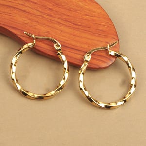 Peut inclure: Une paire de boucles d'oreilles créoles dorées avec un design torsadé. Les boucles d'oreilles sont faites d'un matériau métallique brillant et ont une forme circulaire classique. Les boucles d'oreilles sont présentées sur une surface en bois.