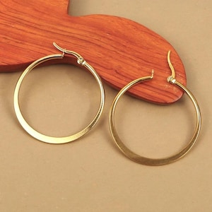 Peut inclure: Deux boucles d'oreilles créoles dorées. Les boucles d'oreilles sont circulaires avec un design simple et poli. Les boucles d'oreilles sont présentées sur une surface en bois avec un fond marron clair.