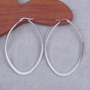 Peut inclure: Une paire de grandes boucles d'oreilles créoles ovales en argent. Les boucles d'oreilles ont une finition lisse et polie et un design simple et élégant. Elles sont présentées sur une surface en bois avec un fond en tissu gris.