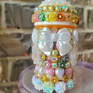 May include: A cylindrical container decorated with a large white bow, iridescent heart-shaped beads, colourful flower-shaped charms, and sparkling rhinestones. The top features a white ruffled cap, and the base is adorned with small pastel floral accents.