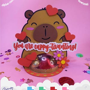 May include: A brown and tan papercraft capybara with red heart accents and the phrase "You are cappy tivanting!" on a white banner. The craft includes a clear container with chocolates and decorative flowers. Digital files are available for Cricut and Silhouette.